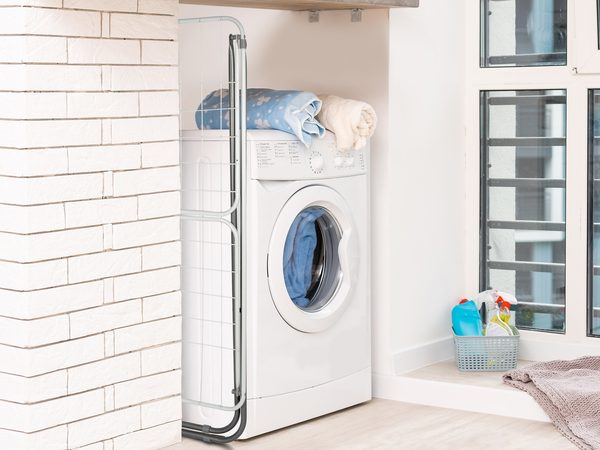 Clothes airers folded flat for storage: a winged airer tucked beside a washing machine and a black 3-tier airer slid behind a sofa
